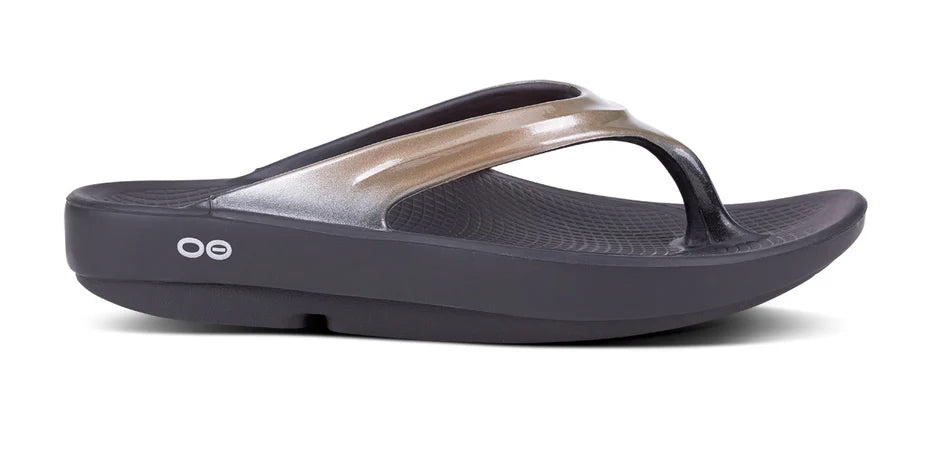 The OOFOS OOlala Luxe Sandal for women is a single black and metallic silver sandal featuring OOfoam technology, a thick sole, textured footbed, and two small white circles on the side.