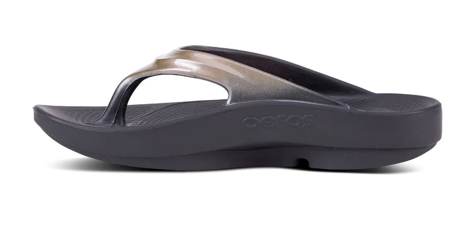 A single OOFOS OOlala Luxe Sandal for women, in black and metallic gray, features a thick OOfoam sole and contoured footbed, shown from the side on a white background.