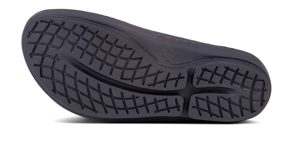 The image shows the black rubber sole of the OOFOS OOlala Luxe Sandal - Women, featuring a wavy tread and crosshatch grooves for grip. OOfoam technology boosts comfort, with embossed brand name and size details on the inner side.