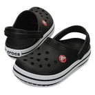 The Crocs CrocBand Clog - Unisex features a black design with white soles, ventilation holes, red circular logos, and an adjustable heel strap, all in a water-friendly, lightweight build. One clog stands upright while the other is tilted.