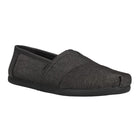The TOMS Classic Canvas - Men is a black slip-on shoe with a classic alpargata design, minimal stitching, no visible laces, textured fabric, flat rubber sole, and a slightly pointed toe for casual comfort.