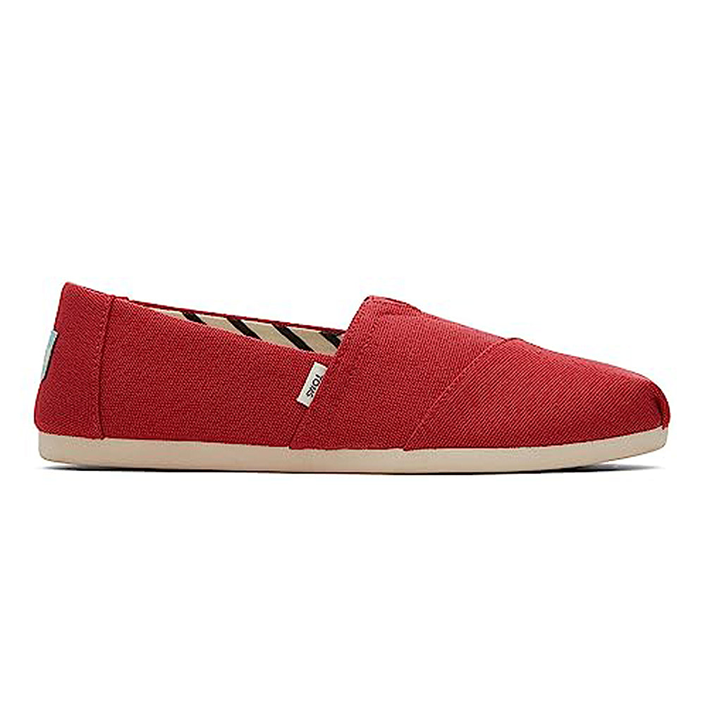 A red canvas TOMS Alpargata ESP slip-on for women with a beige rubber sole, seen from the side. Features a small fabric tag, visible stitching, and highlights TOMS buy one give one commitment.