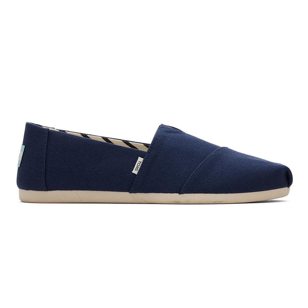 The TOMS Alpargata ESP - Women features a navy blue canvas slip-on design with a beige sole and side fabric tag, shown from the side against a white background—delivering classic TOMS style and signature comfort.