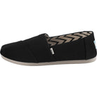 The Alpargata - Womens by TOMS, shown from the side, is a black slip-on from the earthwise collection with recycled cotton canvas, a beige sole, and a chevron-patterned interior lining.