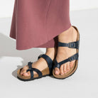A person in a mauve skirt stands on a light surface, wearing black Birkenstock Mayari Oiled Leather sandals featuring cork soles, contoured footbed, and buckles.