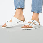 A woman in light blue jeans and chunky double strap Birkenstock Arizona sandals with silver buckles stands on a white surface, highlighting orthopedic walking comfort in this classic design.