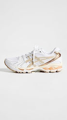 The ASICS Gel-Kayano 14 - Womens sneaker features a white mesh and synthetic build, metallic gold accents, GEL Technology for comfort, a chunky sole, and a lace-up design.