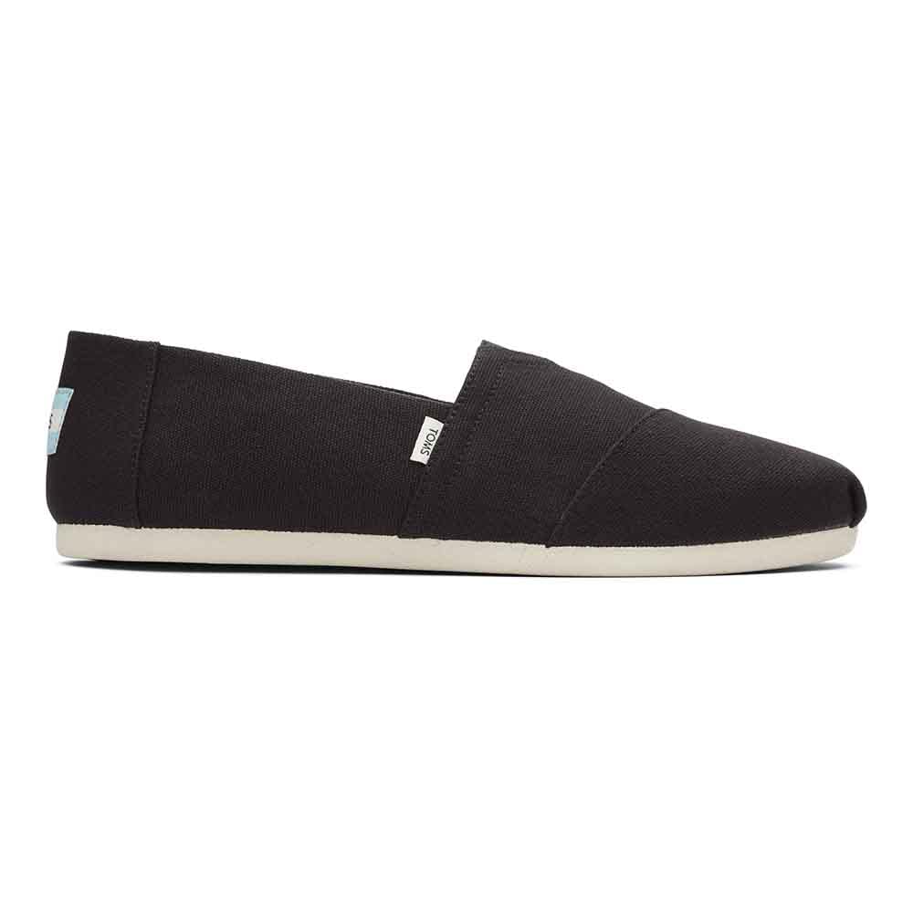 The Classic Canvas - Men by TOMS is a black slip-on shoe with a white sole, shown in side view. Its minimalist design includes small fabric tags near the heel and side seam, reflecting the classic TOMS look.