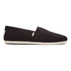 The Classic Canvas - Men by TOMS is a black slip-on shoe with a white sole, shown in side view. Its minimalist design includes small fabric tags near the heel and side seam, reflecting the classic TOMS look.