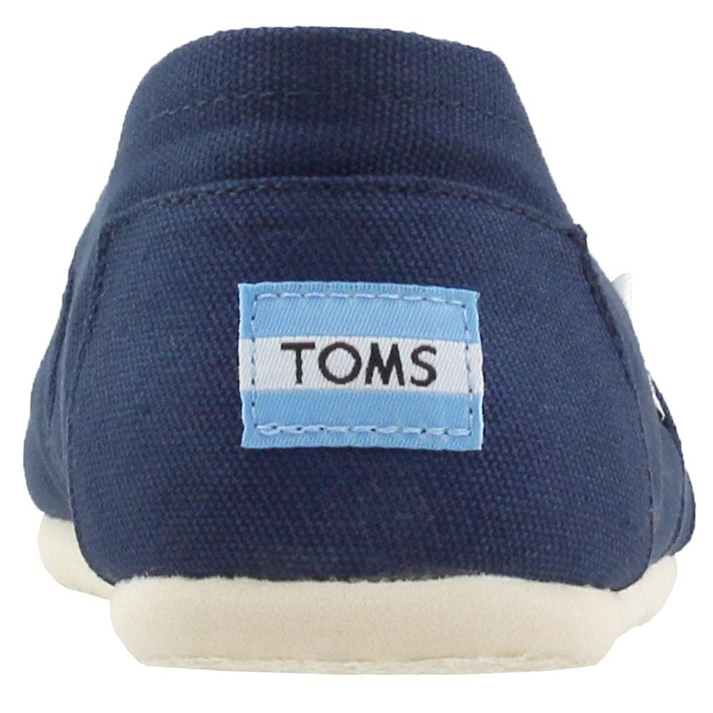 The image shows the back of a TOMS Classic Canvas - Men navy blue slip-on with a light blue and white “TOMS” logo patch on the heel and an off-white sole.