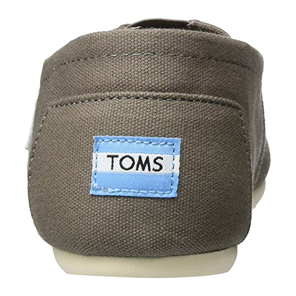 A close-up rear view of the TOMS Classic Canvas - Men shoe in brown, showing a blue and white TOMS label on the heel, off-white rubber soles, and a classic slip-on design.