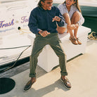 A man and woman smile on a dock by the water, both in classic Sperry shoes. He wears Sperry A/O 2-Eye Leather – Mens with a navy shirt and olive pants; she pairs hers with stripes and shorts, enjoying a sunny day.