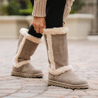 Wearing tall, tan BEARPAW Kendall Boots lined with white faux fur, a person dressed in black leggings and a beige sweater bends down on a paved outdoor surface near Lake Tahoe.