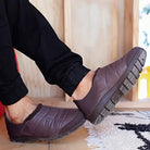 A person wears BEARPAW Harry Slippers - Mens, featuring thick rugged soles, standing indoors on a light floor near a rug and wooden wall—highlighting these cozy slippers designed for mens comfort.