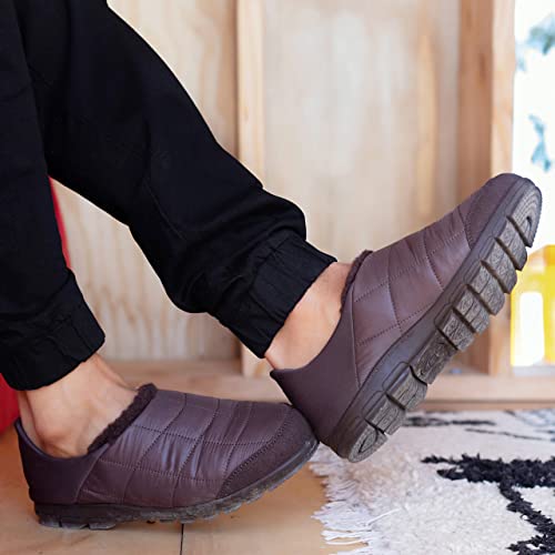 A person wears BEARPAW Harry Slippers - Mens, featuring thick rugged soles, standing indoors on a light floor near a rug and wooden wall—highlighting these cozy slippers designed for mens comfort.