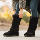 A woman in blue jeans and a checked coat adjusts her tall, black, fur-lined BEARPAW Kendall Boots outdoors on a concrete path.