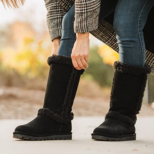 A woman in blue jeans and a checked coat adjusts her tall, black, fur-lined BEARPAW Kendall Boots outdoors on a concrete path.