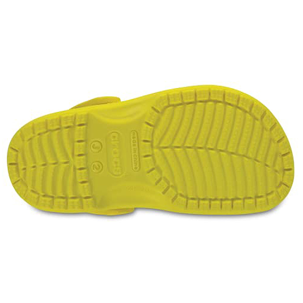 The image shows the yellow sole of a Crocs Classic Clog - Kids shoe, viewed from the bottom, highlighting its tread pattern and the Crocs logo in the center.