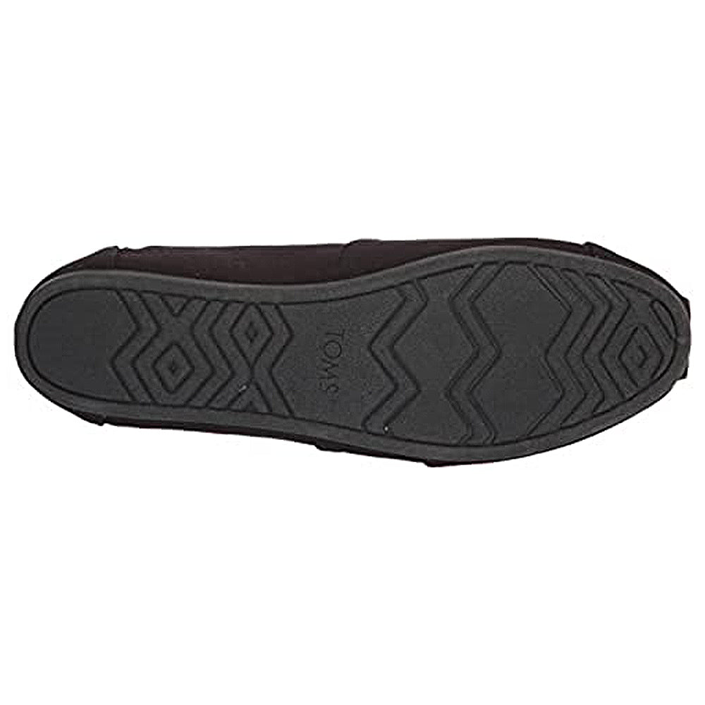 The image shows the bottom view of a black TOMS Alpargata - Womens shoe, featuring a textured rubber sole with zigzag and diamond patterns and the TOMS name embossed in the center.