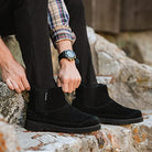 A man wearing BEARPAW Nick Boots and black pants sits on rocks, adjusting a boot. His look is completed with a plaid shirt and a sleek black wristwatch.