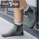 A person in tan pants and gray Hey Dude Scott Grip Boots stands on a sidewalk by a glass wall, highlighting these mens weather-ready Chelsea boots with ultra grip outsoles for stability.