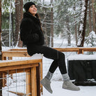 A woman wearing BEARPAW Shorty Boots sits on a snowy wooden fence in a forest, dressed in a black jacket, black leggings, and a black beanie, smiling as she looks up.