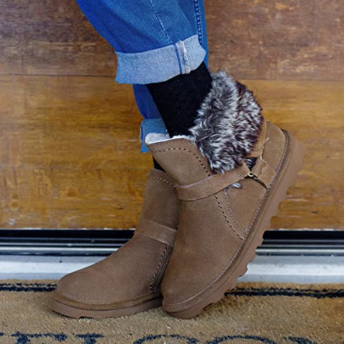 A woman wearing blue jeans and black socks stands indoors against a wooden door, modeling BEARPAW Konnie Boots - Womens with faux fur lining and textured sole.