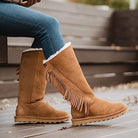 A person in blue jeans and tan BEARPAW Tamara Boots for women with fringe detailing sits on wooden steps outdoors.