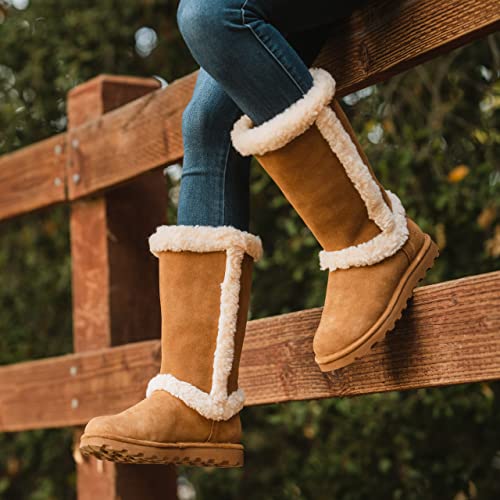 A woman in blue jeans and tan, fur-lined BEARPAW Kendall Boots sits on a wooden fence surrounded by green foliage, capturing cozy Lake Tahoe vibes.