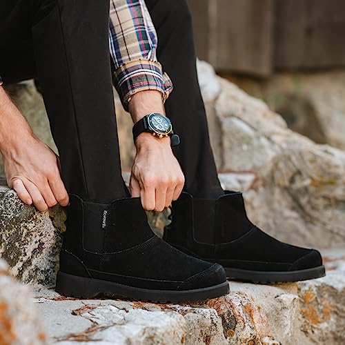 A person sits on rocks adjusting their black pants, revealing chic BEARPAW Nick Boots for men. They wear a plaid shirt and a wristwatch.
