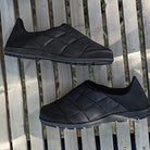 A pair of BEARPAW Harry Slippers - Mens in black, featuring quilted uppers and rubber soles, displayed on a wooden slatted surface and viewed from above.