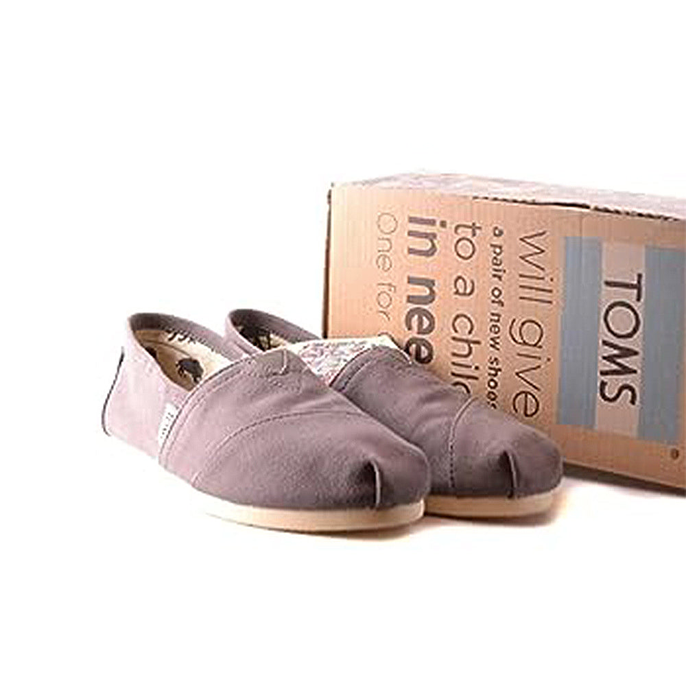 A pair of TOMS Classic Canvas - Men gray slip-on shoes sits before a cardboard box with the TOMS logo and a message about donating shoes to children in need, against a white background.