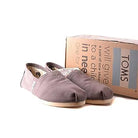 A pair of TOMS Classic Canvas - Men gray slip-on shoes sits before a cardboard box with the TOMS logo and a message about donating shoes to children in need, against a white background.
