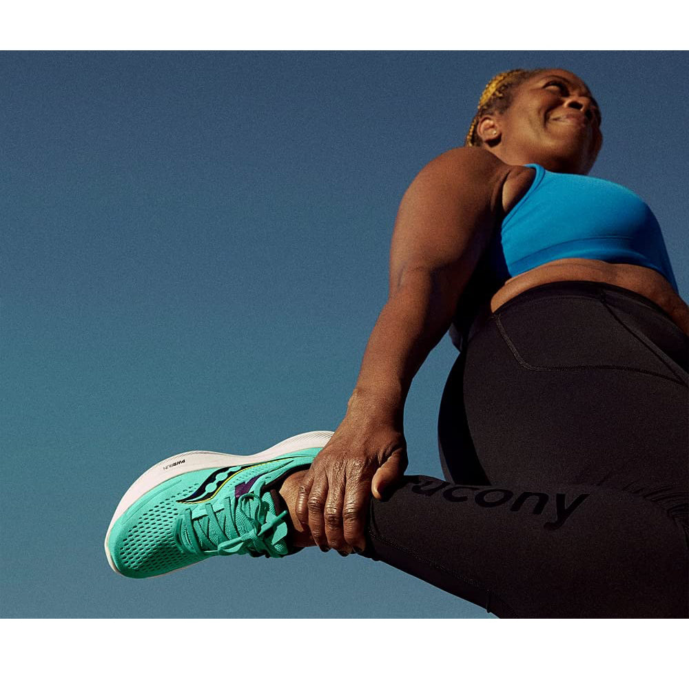 A woman in a blue sports bra and black leggings stretches her leg, holding her turquoise Saucony Ride 15 - Womens running shoe with PWRRUN cushioning against a clear blue sky.