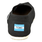 A back view of the TOMS Classic Canvas - Men slip-on in black, showing a blue and white rectangular TOMS logo tag above a white sole.