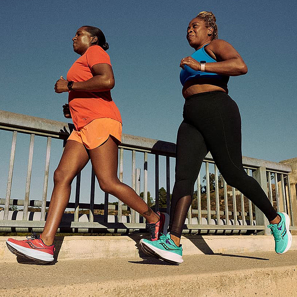Two women jog together on a sunny day—one in orange, the other in a blue top and black leggings—enjoying the comfort of Saucony Ride 15 - Womens shoes as they run along a path by a metal railing.