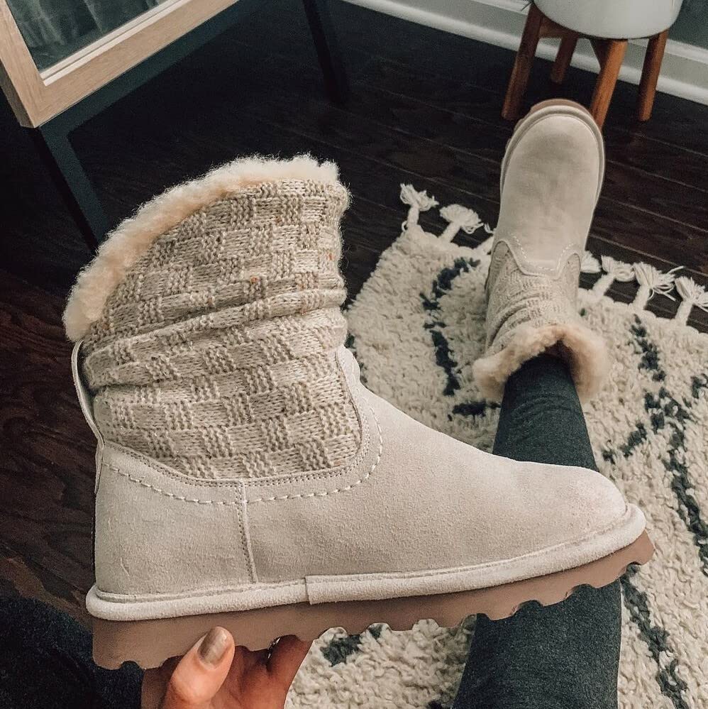 A person in dark leggings holds a BEARPAW Virginia Boot—womens beige suede and wool knit shaft winter boot with fur lining—while wearing the matching sheepskin boot on one foot, standing on a patterned rug indoors.