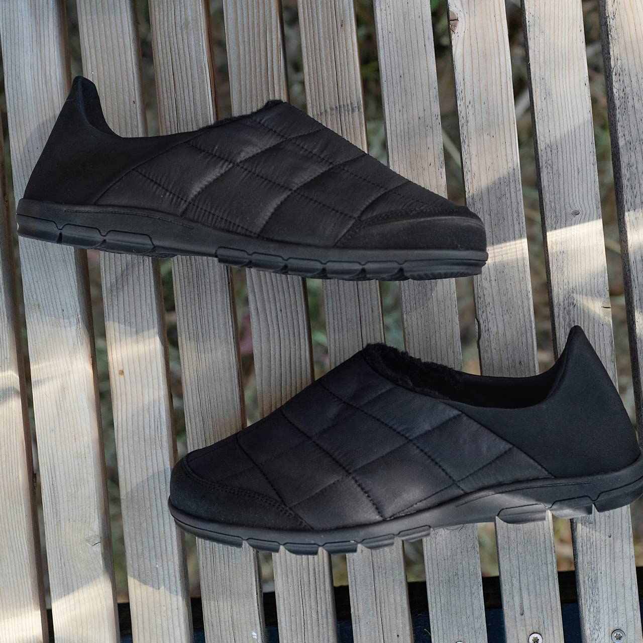 A pair of BEARPAW Harry Slippers for men with quilted uppers and rubber soles rests on a wooden slatted surface, viewed from above.