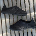 A pair of BEARPAW Harry Slippers for men with quilted uppers and rubber soles rests on a wooden slatted surface, viewed from above.