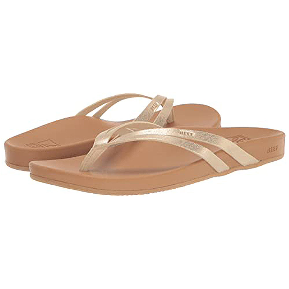The Reef Cushion Spring Joy womens flip flops feature tan soles, metallic gold straps, and a cushioned footbed for comfort, shown against a white background. Their simple, casual design is ideal for everyday wear.