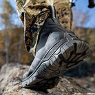 Close-up of a person wearing the Apex Mid Zip - Men rugged outdoor boots by The Original Muck Boot Company with camouflage pants, stepping on rocky terrain outdoors against a blurred background of trees.