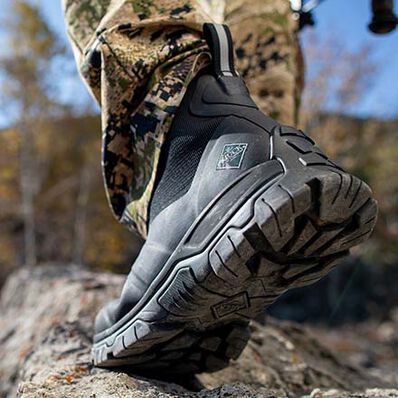 Close-up of a person wearing the Apex Mid Zip - Men rugged outdoor boots by The Original Muck Boot Company with camouflage pants, stepping on rocky terrain outdoors against a blurred background of trees.