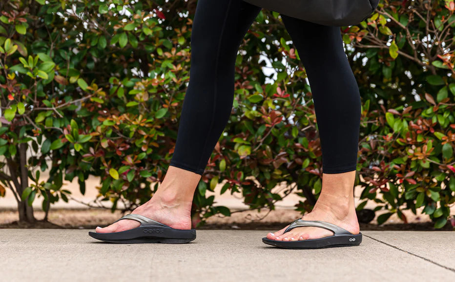 A person in black leggings and OOFOS OOlala Luxe Sandals, featuring OOfoam technology, walks on a sidewalk with green and reddish bushes in the background.