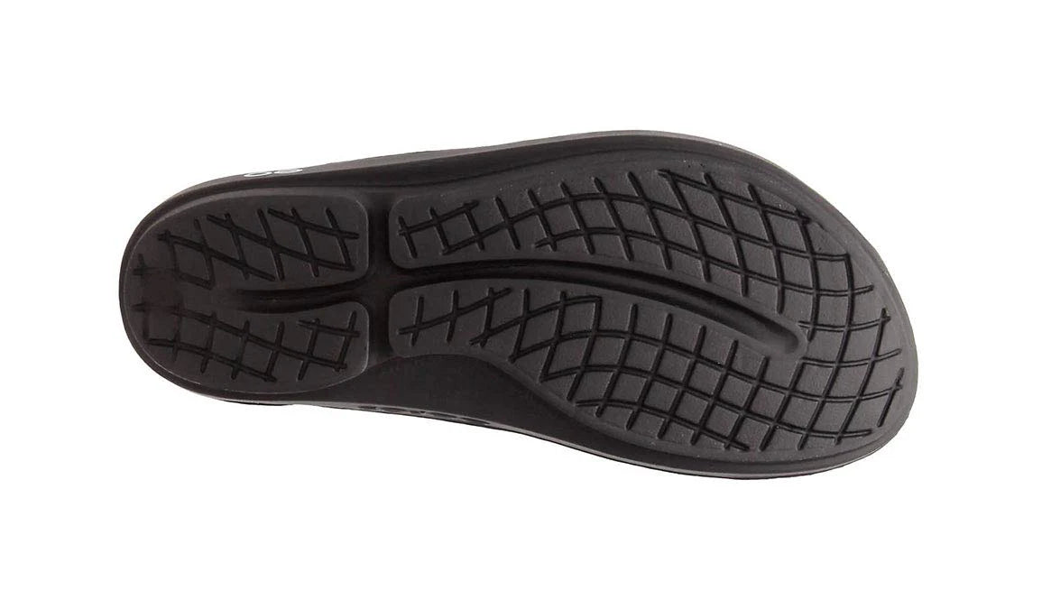 The image shows the black rubber sole of the OOFOS OOlala Sandal - Unisex, featuring a curved tread pattern and crisscrossed grooves for traction with OOfoam technology, viewed from the bottom against a white background.